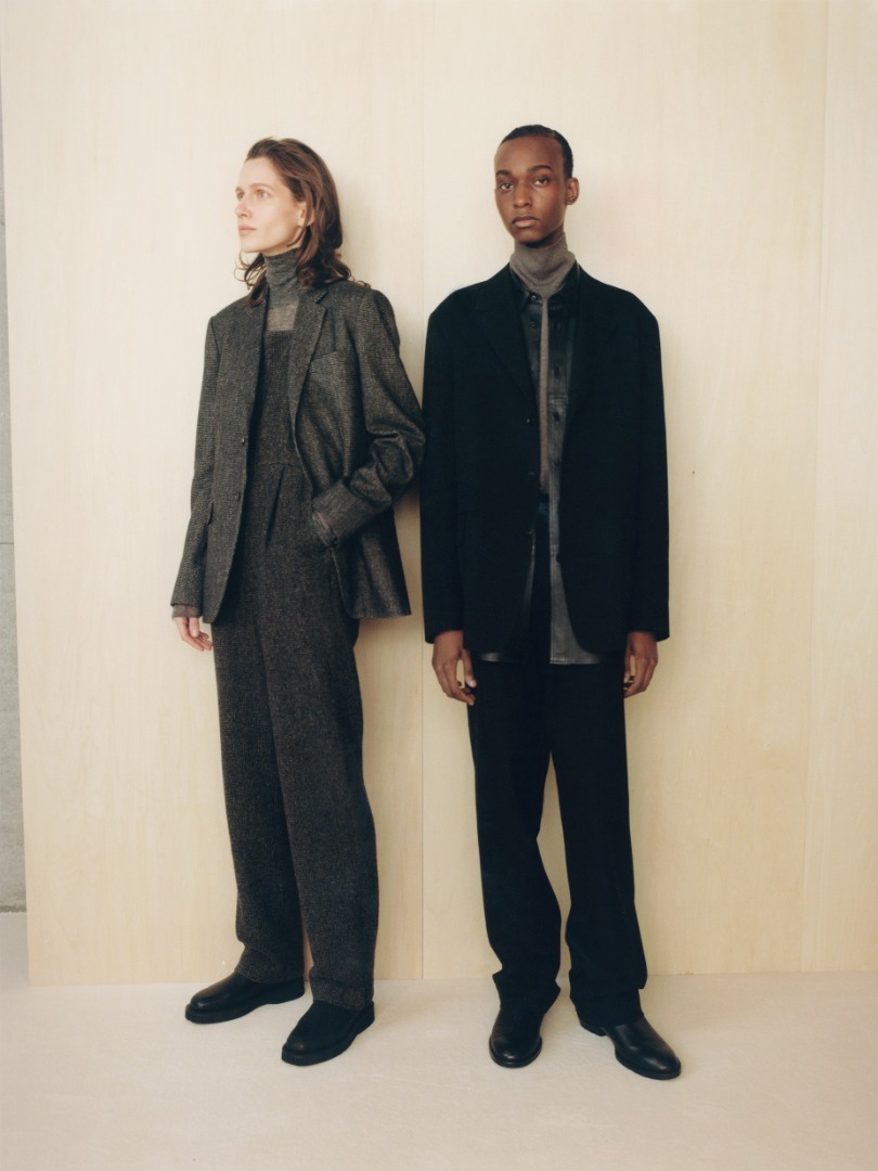Ella wears Organic Cotton Cashmere Wool Tweed Jacket in Top Charcoal, Wool Hairline Light Tweed Overalls in Top Charcoal. Mohamed wears Super Fine Wool Cotton Twill Over Jacket in Black, Super Fine Wool Cotton Twill Slacks in Black